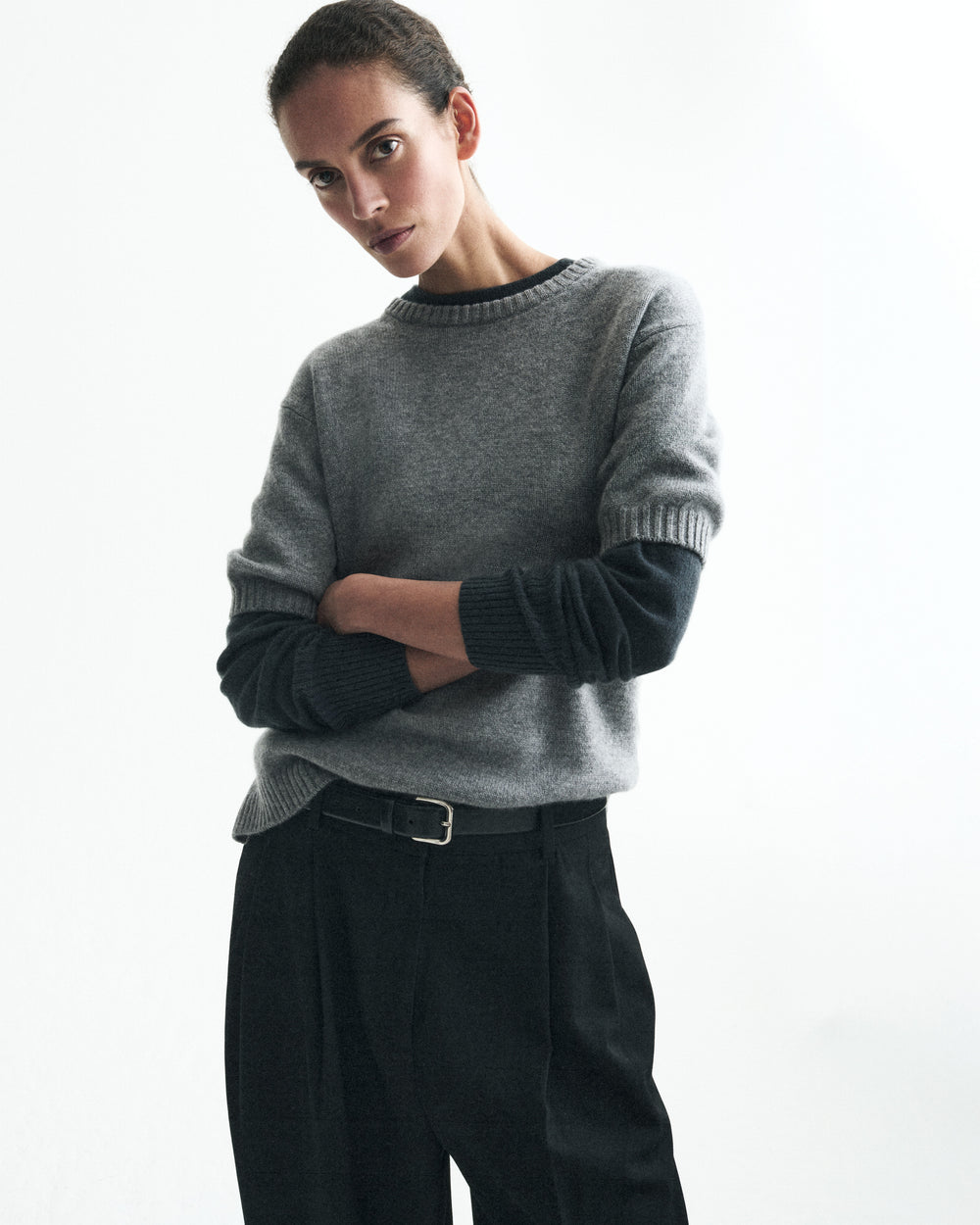 A model wearing a gray crewneck sweater with contrasting dark charcoal ribbed trim on the collar, cuffs, and sleeves, paired with black trousers, against a clean white studio background.