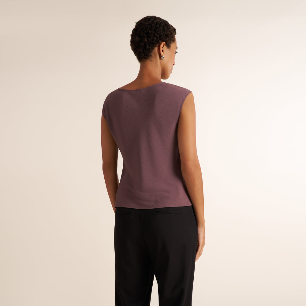 A woman shown from the back wearing a sleeveless mauve top and black pants, standing against a plain light beige studio background.