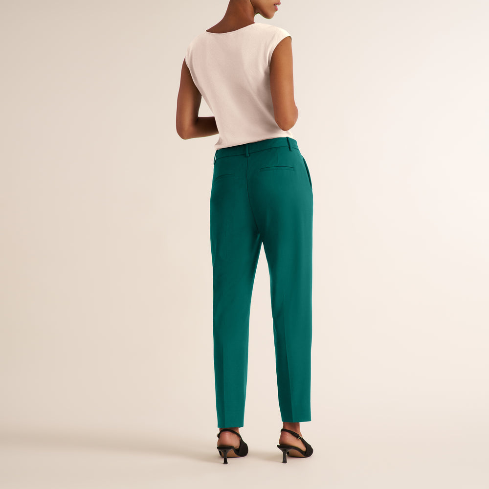 A model shown from the back wearing high-waisted teal trousers, a white sleeveless top, and black heeled sandals against a plain beige studio background.