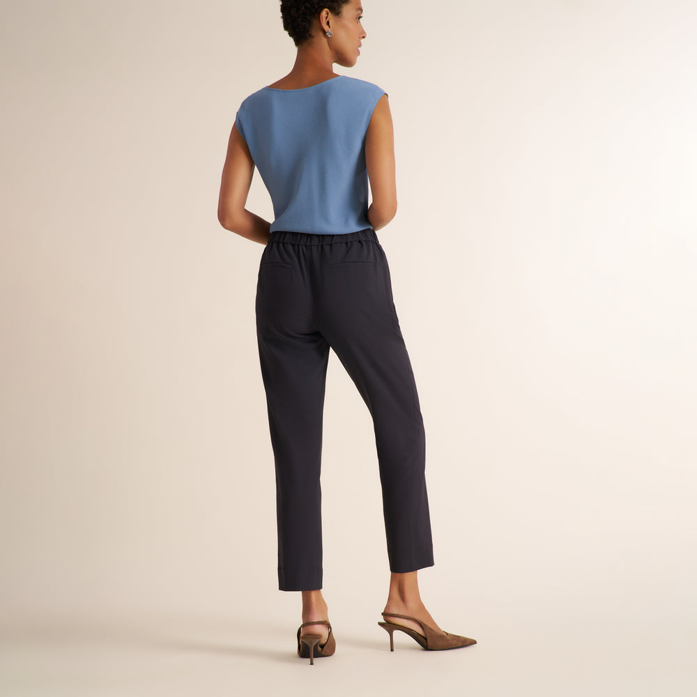 A model stands against a warm off-white studio background wearing a light blue sleeveless top with a draped side detail and dark navy cropped trousers, paired with brown pointed-toe heels.
