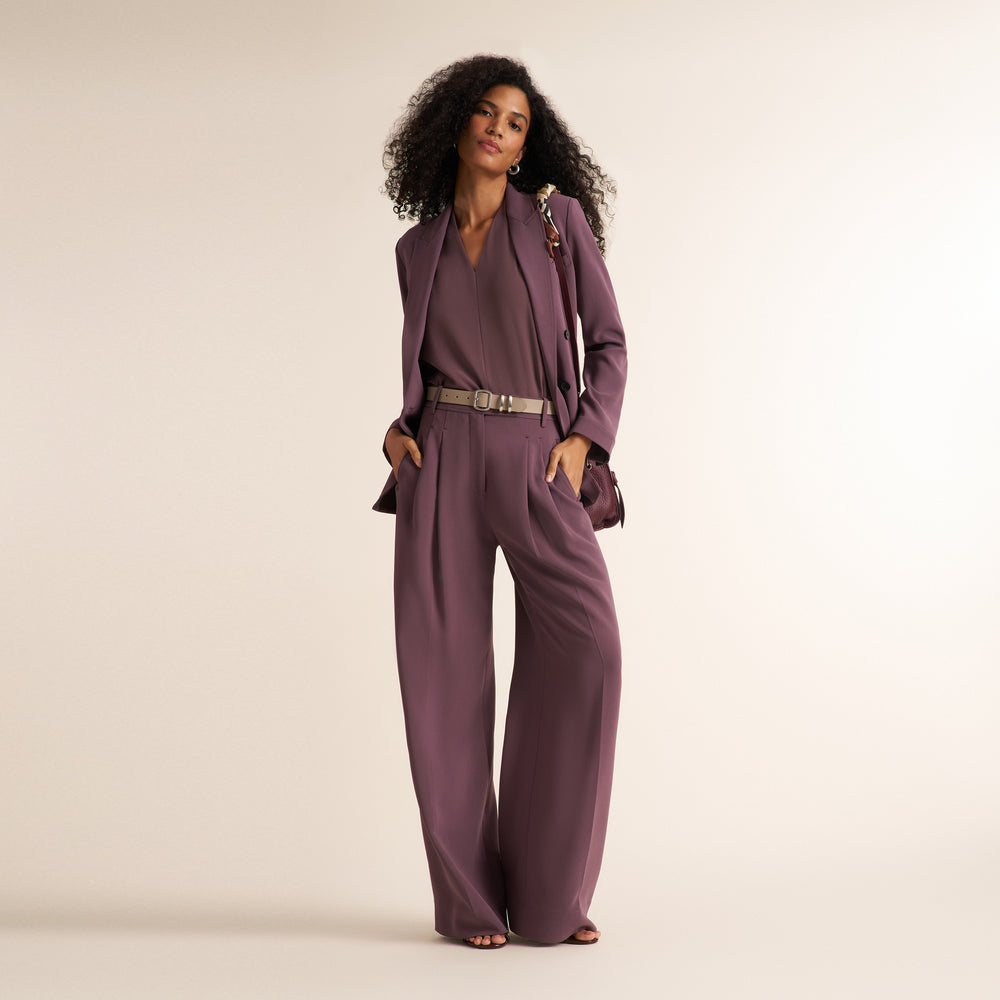 A woman modeling a matching mauve-purple two-piece set consisting of a V-neck jumpsuit and a tailored blazer, styled with a silver belt and a small matching handbag against a solid cream background.