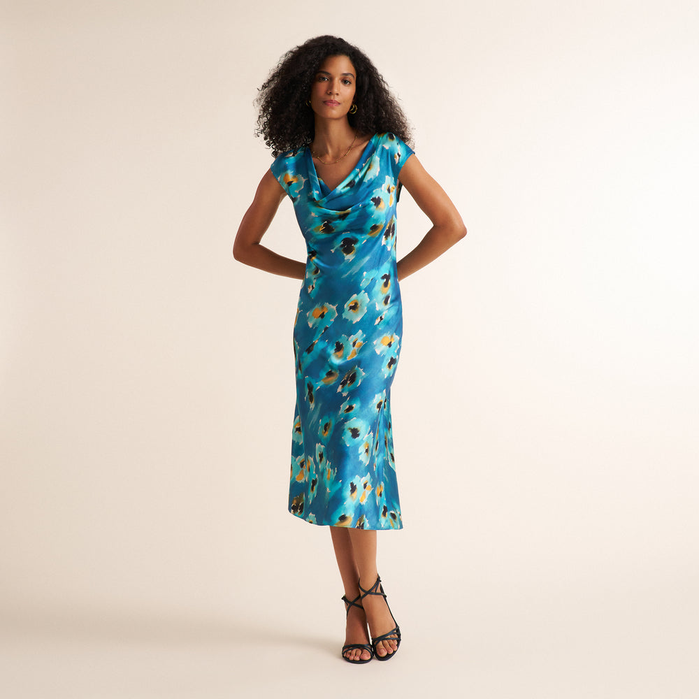 A woman modeling a fitted cowl-neck midi dress with a blue base and abstract floral print in white, yellow, and black, paired with black strappy heels against a plain beige studio background.