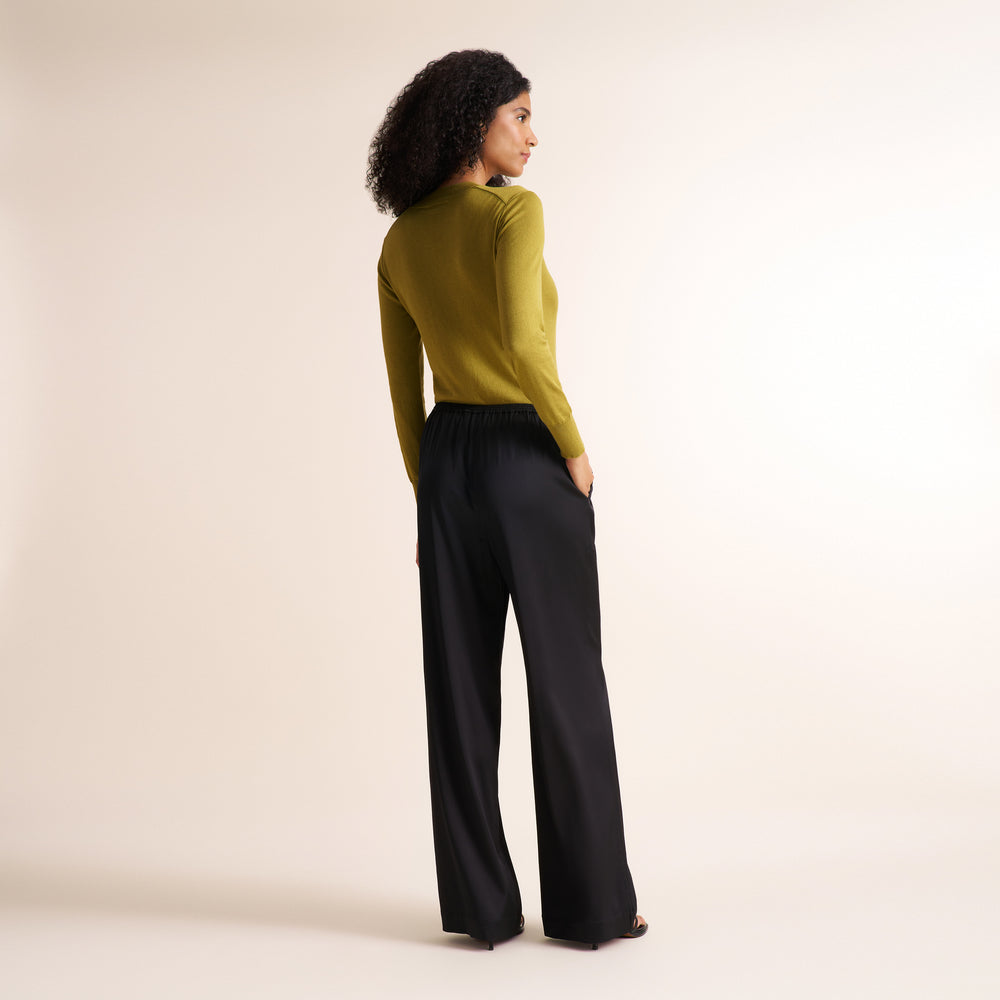 A woman with curly black hair is shown from a side-back angle wearing a fitted olive green long-sleeve top and black wide-leg pants against a plain warm off-white studio background.
