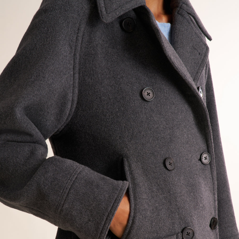 A close-up of a person wearing a dark charcoal gray double-breasted wool coat with black buttons, showing the collar, lapels, and side pocket.