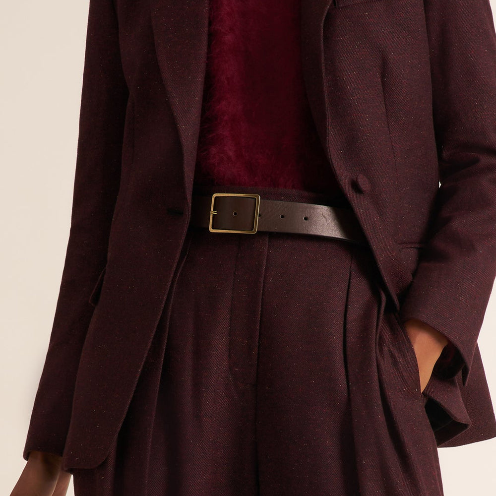 A woman modeling a tailored dark burgundy blazer with matching high-waisted trousers, worn over a textured burgundy top and a dark belt, against a plain light beige studio background.