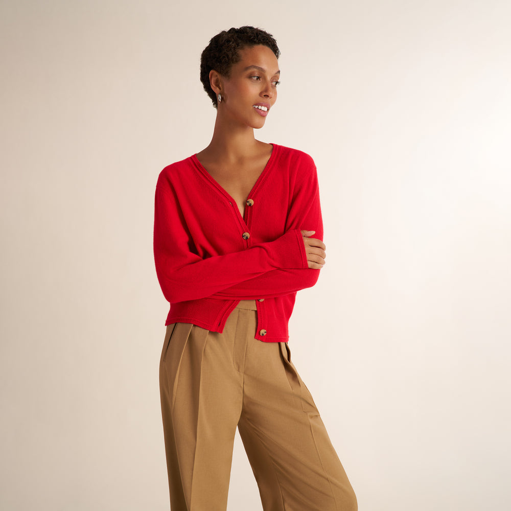 A model wearing a vibrant red knit cardigan with gold buttons and tan trousers, standing against a plain beige studio background.