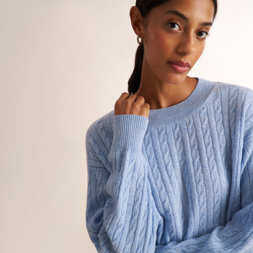 A brunette woman modeling a blue melange long-sleeve knit top with a a crew neck collar and cable knit detail, paired with saddle wide leg trousers, against a plain light beige studio background.