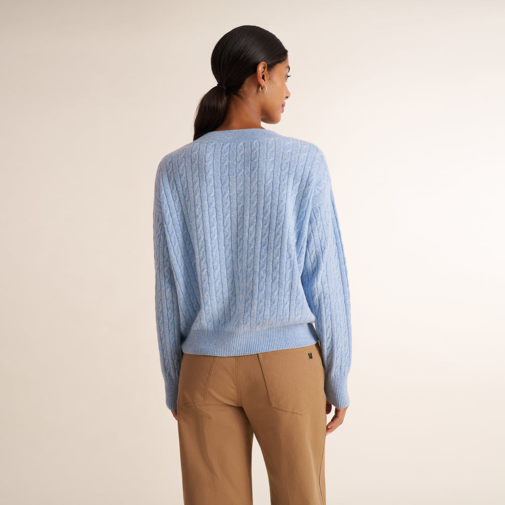 A brunette woman modeling a blue melange long-sleeve knit top with a a crew neck collar and cable knit detail, paired with saddle wide leg trousers, against a plain light beige studio background.