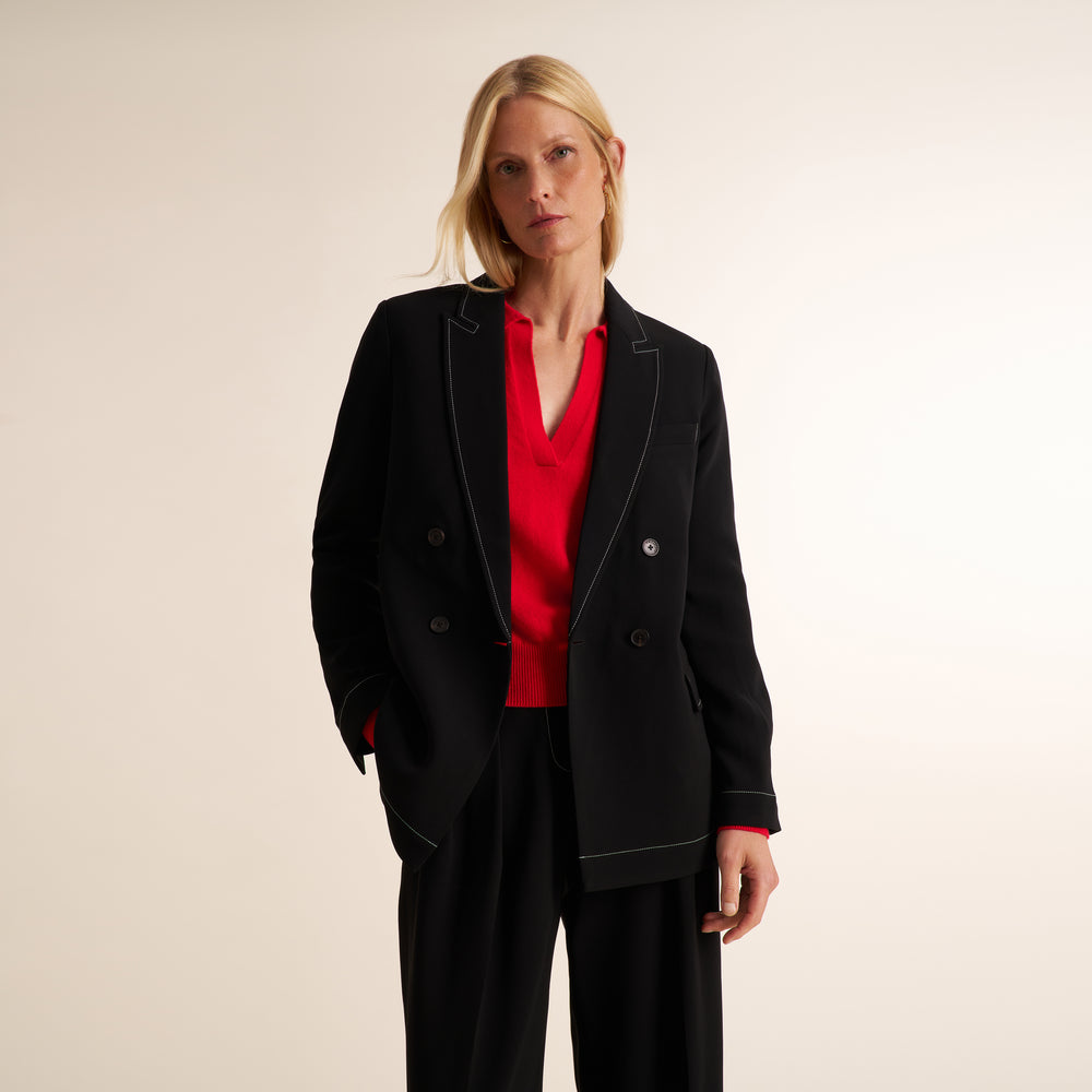 A blonde woman modeling a black double-breasted blazer with white contrast piping over a vibrant red V-neck blouse and black trousers, set against a plain beige studio background.