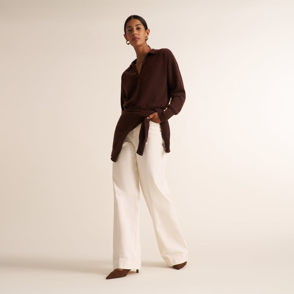 A model stands in a studio wearing a dark brown wrap-style knit top with wide-leg white pants and brown pointed shoes against a warm off-white background.