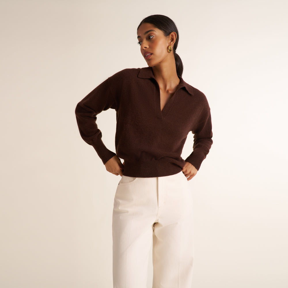 A woman modeling a dark brown knit polo-neck sweater paired with off-white trousers, standing against a plain light beige studio background.