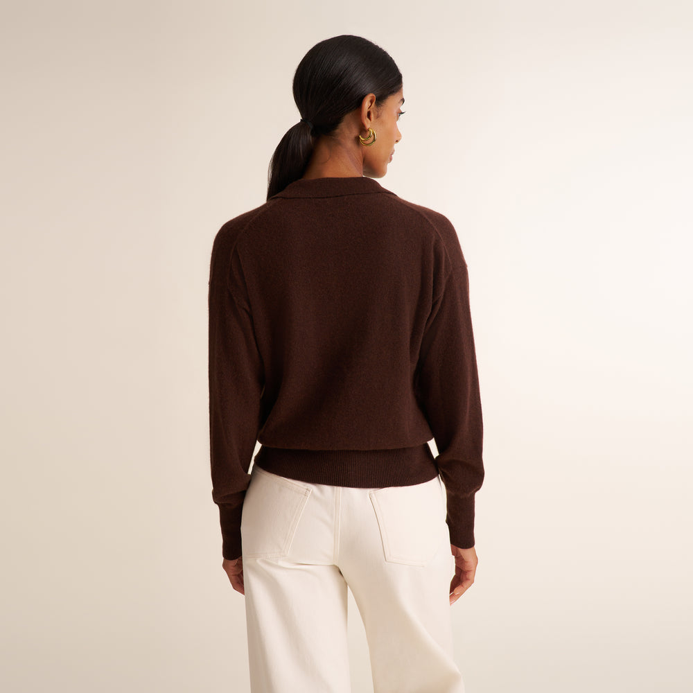 A woman shown from the back wearing a dark brown cropped knit cardigan and off-white trousers, standing against a plain light beige studio background.