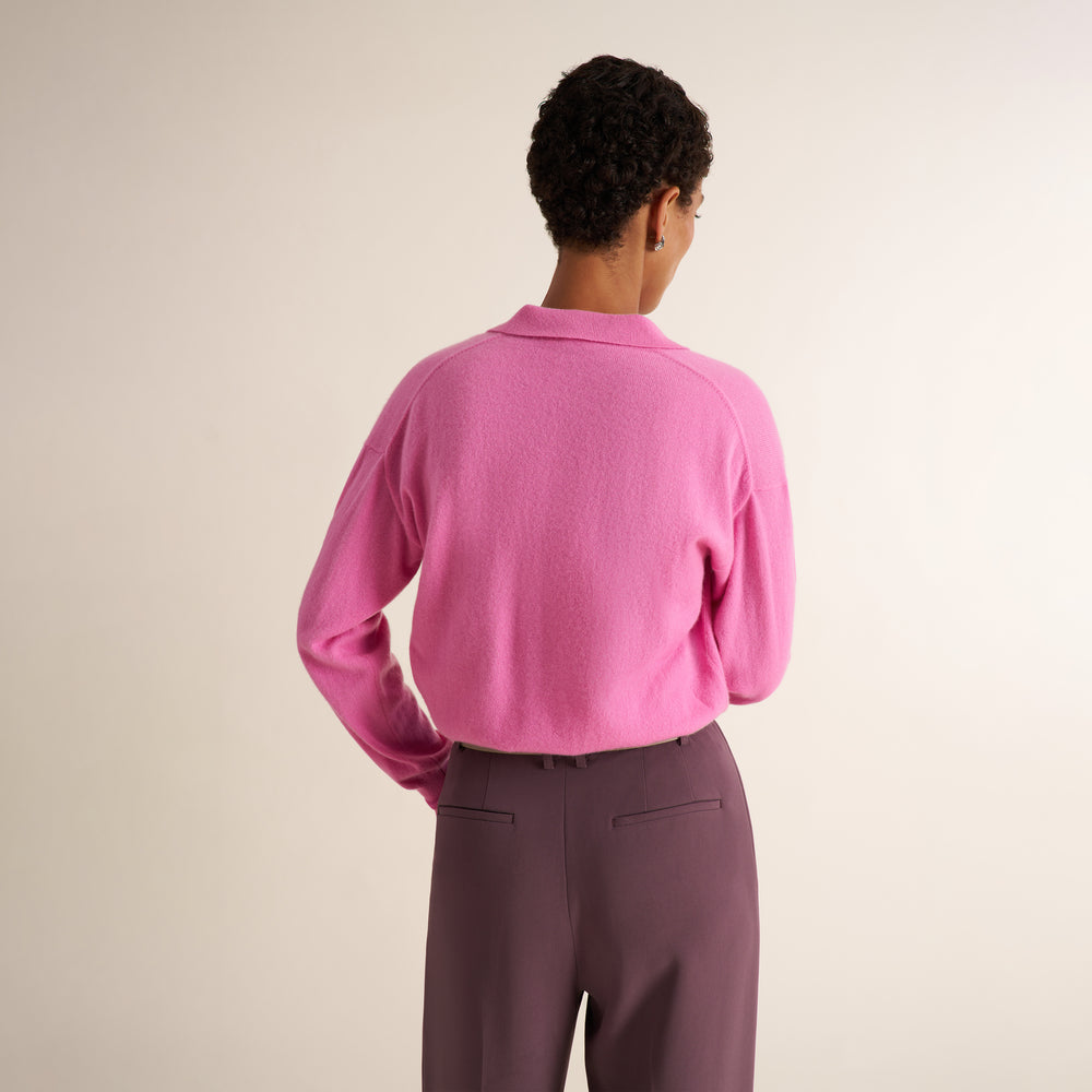 A brunette woman modeling the back of a pink long-sleeve knit top with a split V-neck collar, paired with purple trousers, against a plain light beige studio background.