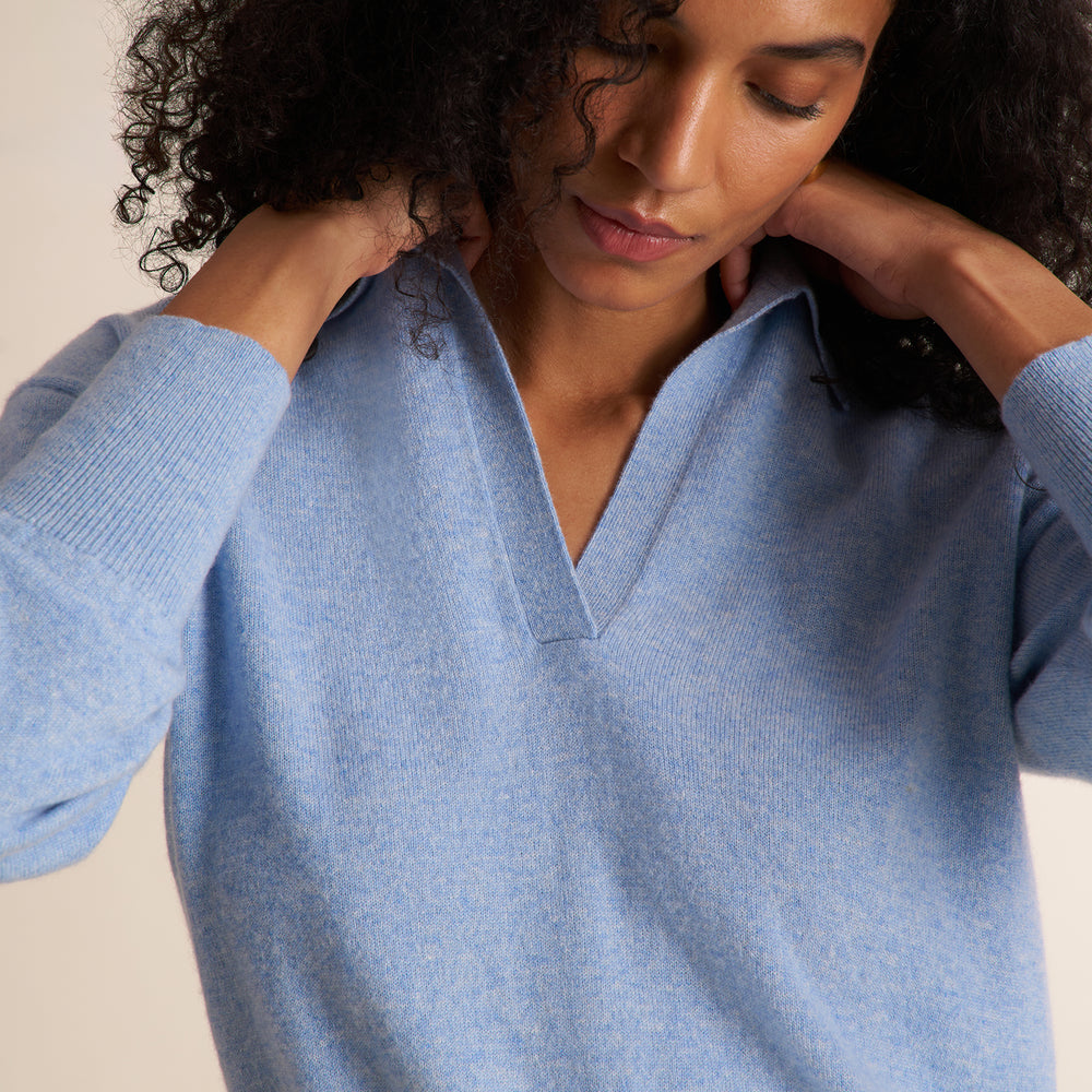 A curly brunette woman modeling a blue melange long-sleeve knit top with a split V-neck collar, paired with ivory wide leg trousers, against a plain light beige studio background.