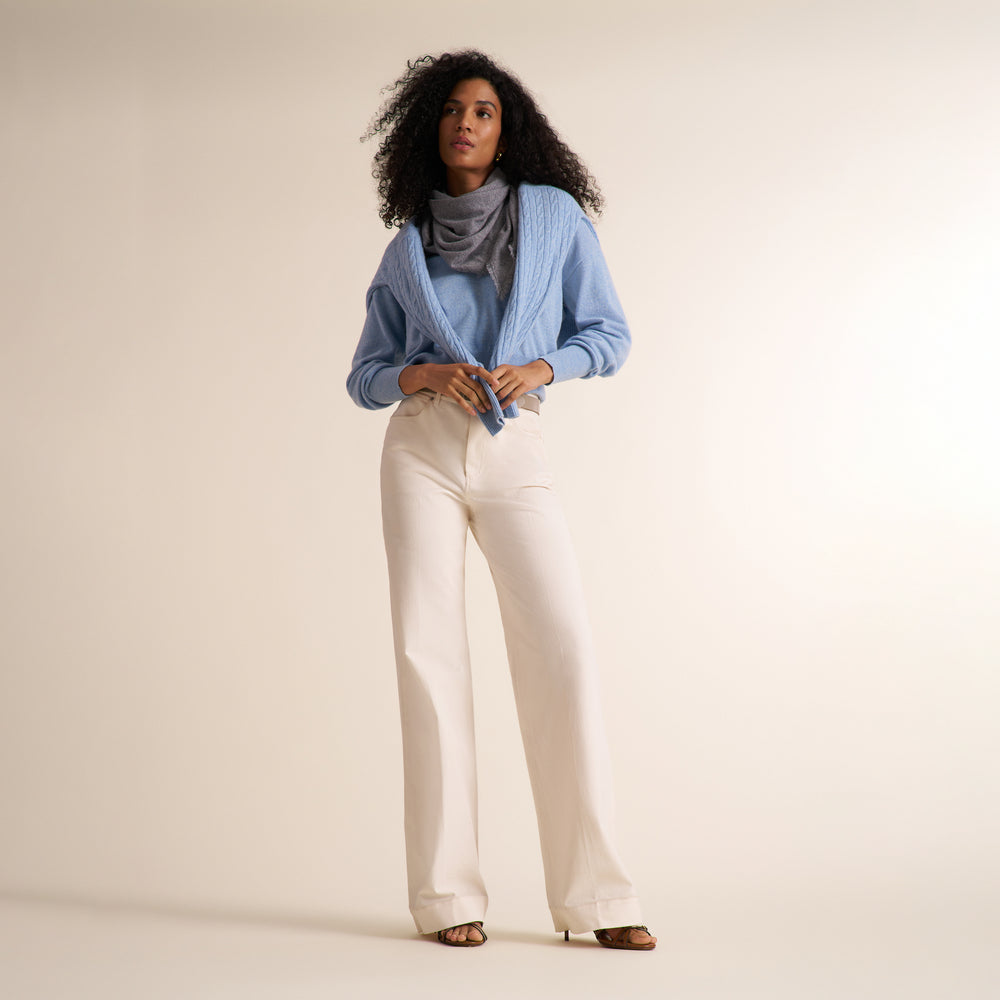 A curly brunette woman modeling a blue melange long-sleeve knit top with a split V-neck collar, paired with ivory wide leg trousers, against a plain light beige studio background.