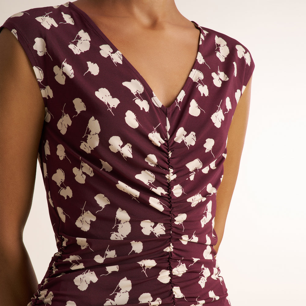 A close-up of a woman wearing a sleeveless V-neck dress in deep burgundy with a white floral print and gathered ruching down the front.