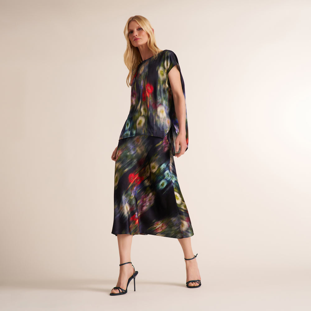 A woman modeling a short-sleeved midi dress with a dark abstract multicolor print, standing against a plain beige studio background.