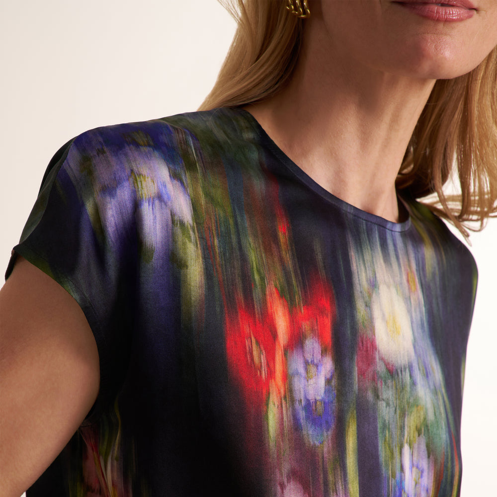 A close-up of a woman wearing a short-sleeve crewneck top with an abstract painterly print in dark blue, purple, green, red, and white.