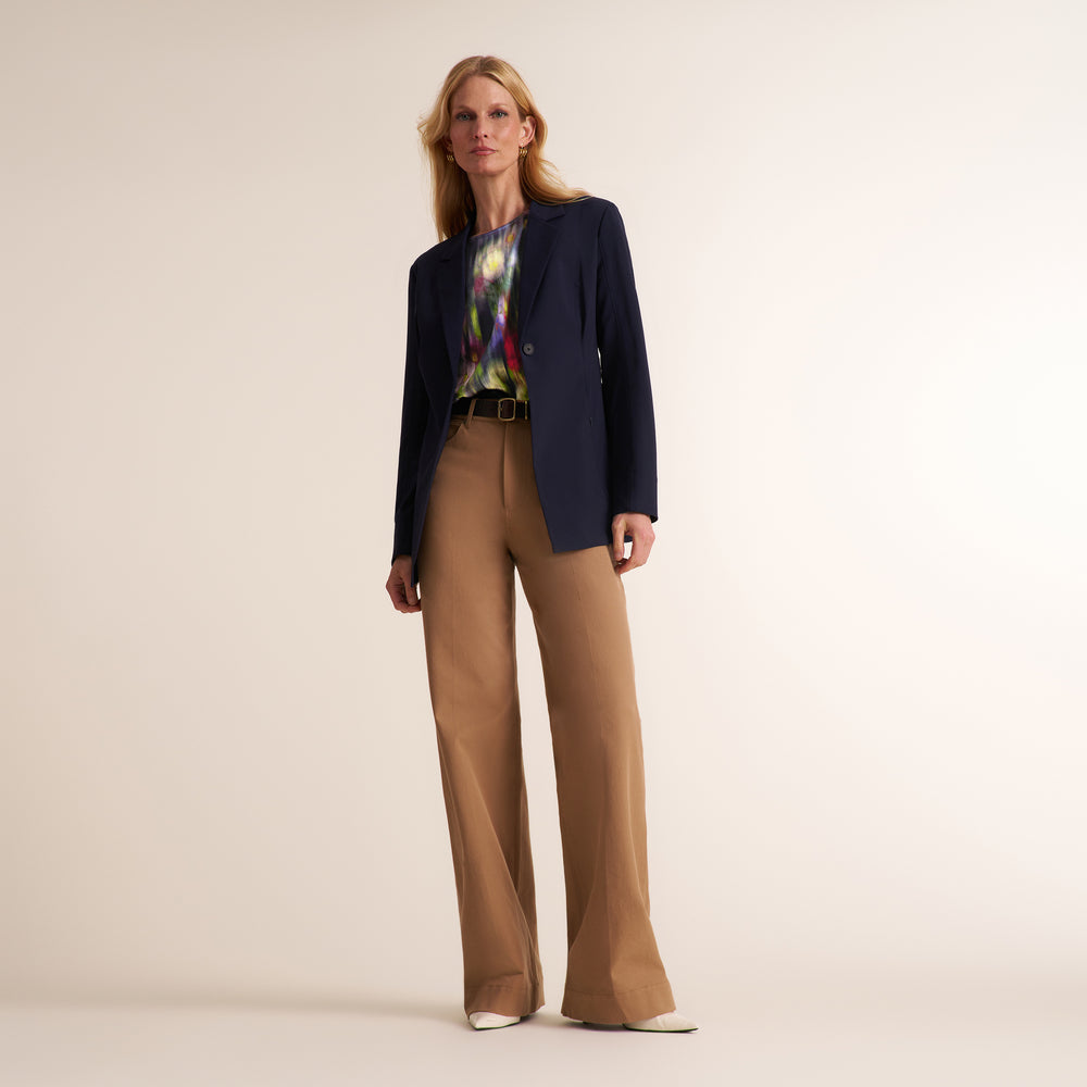 A model stands against a plain light beige studio background wearing a dark navy blazer, wide-leg tan pants, a colorful tie, and white pointed shoes.