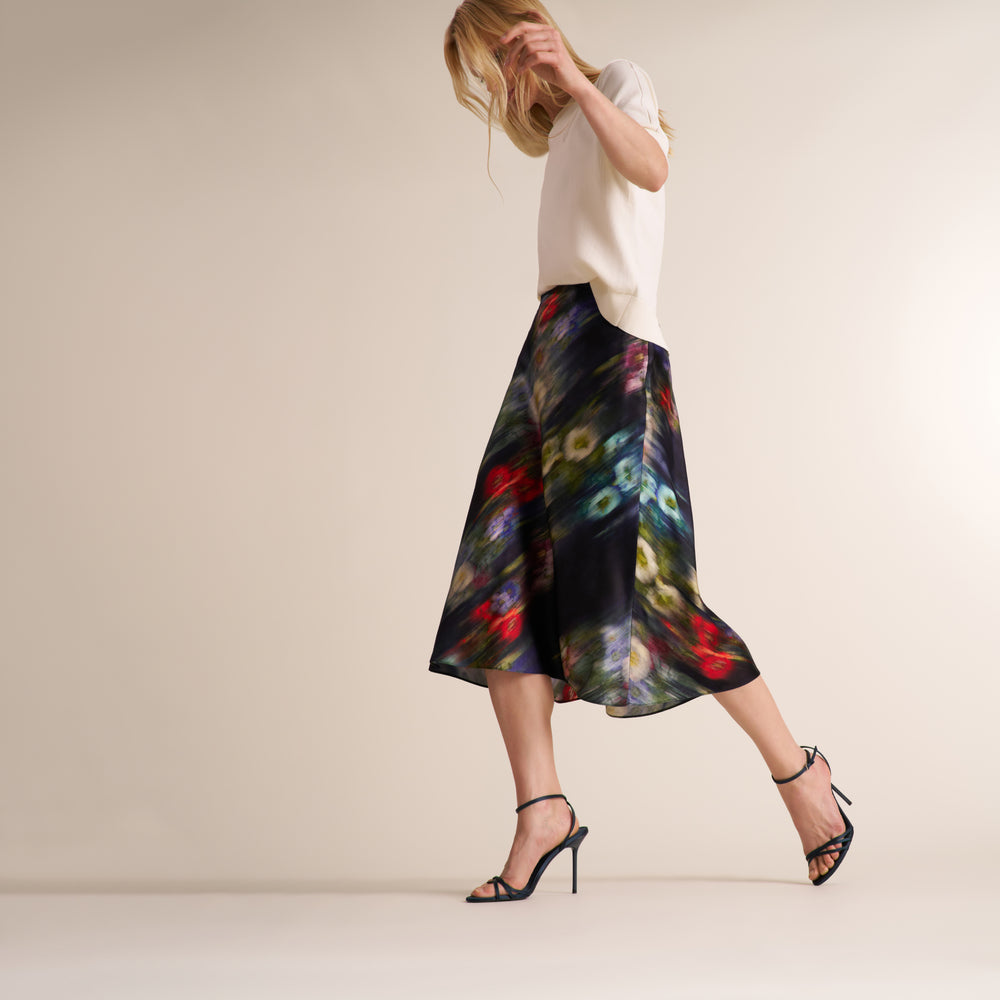 A woman models a black midi skirt with a vibrant, painterly multicolor abstract print, paired with a cream top and black strappy high-heeled sandals, set against a plain beige studio background.