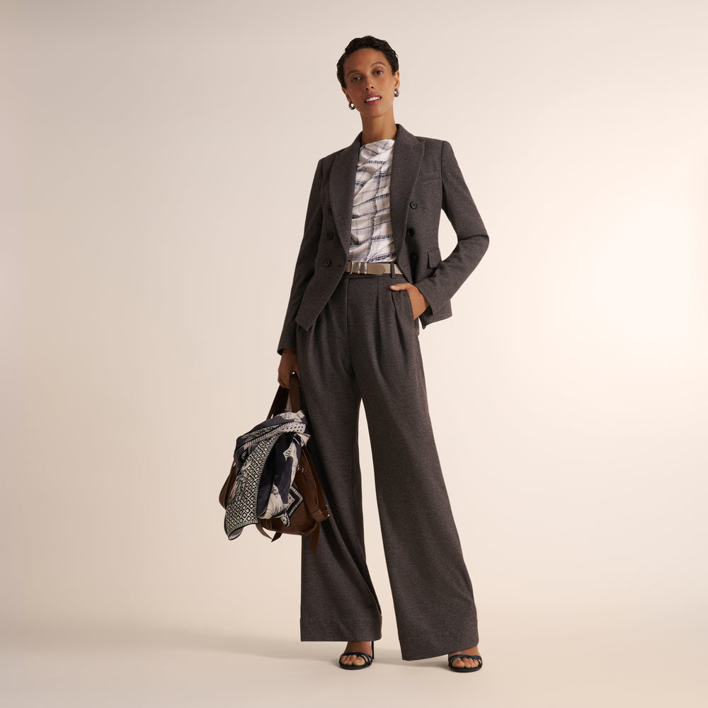 A model stands against a warm beige studio background wearing a dark gray two-piece suit with wide-leg trousers and a matching blazer, layered over a white patterned top. She holds a patterned scarf and a brown leather bag.