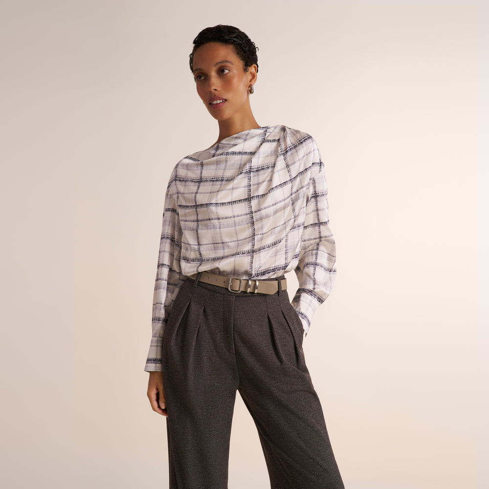 A model wearing a white long-sleeve top with a gray and black plaid pattern, tucked into dark gray pleated trousers with a beige belt, against a solid light beige background.