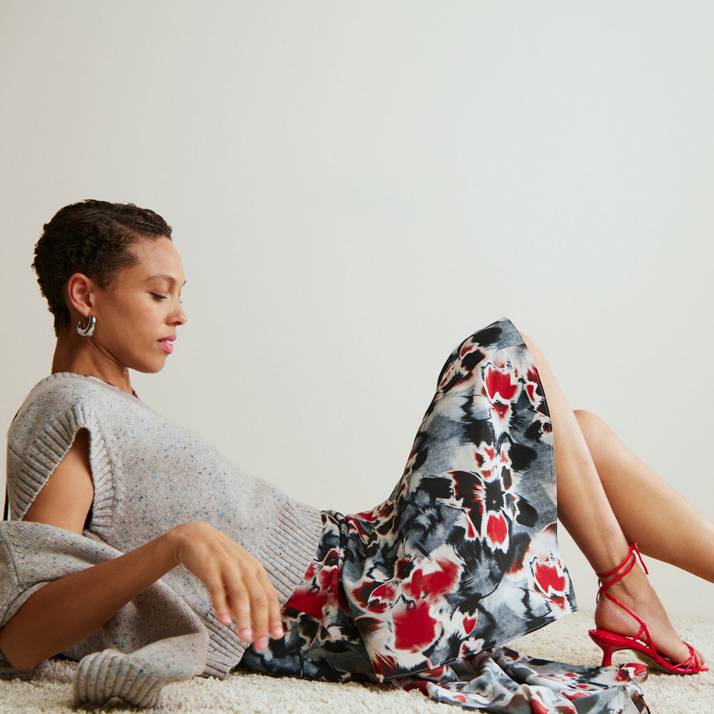 Woman sitting on a light-colored surface wearing a gray sweater and floral skirt with red shoes.
