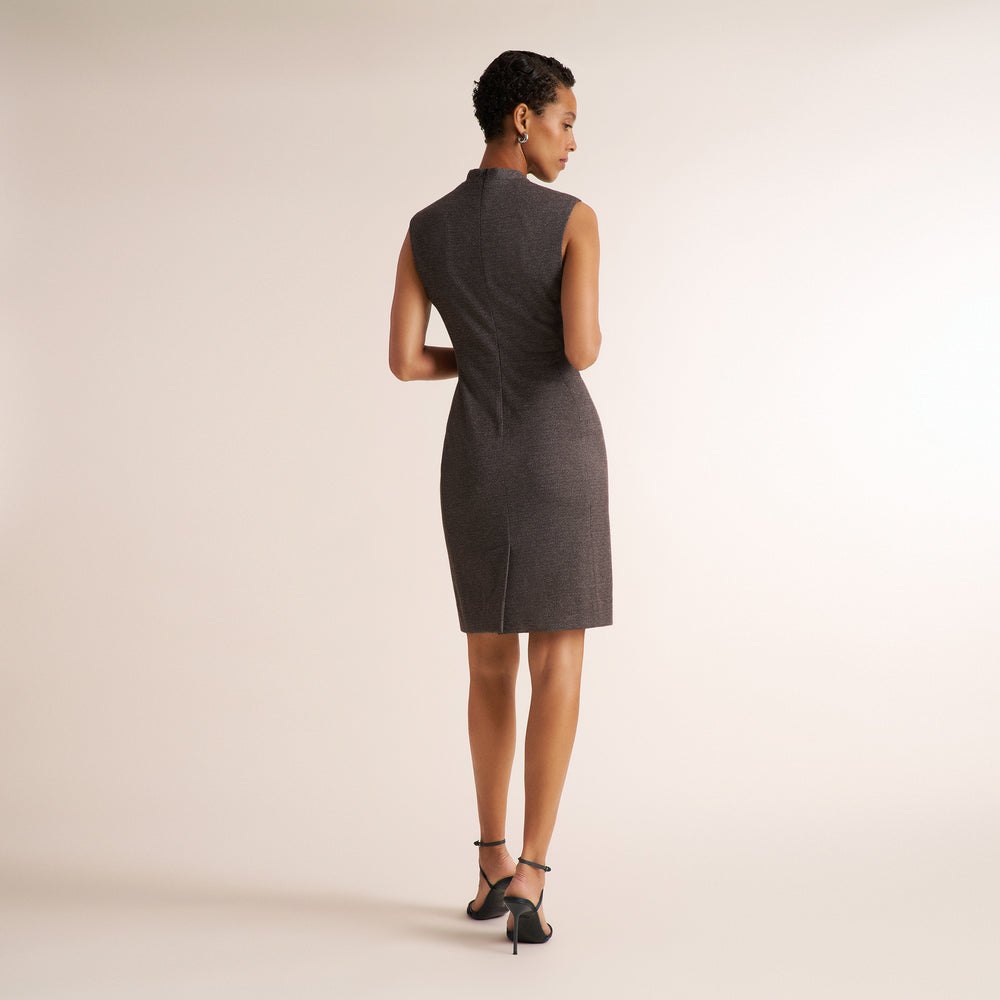 A woman is shown from the back wearing a sleeveless, knee-length dark gray dress with a high mock neck and a small front slit, paired with black strappy heels, against a solid light beige studio background.