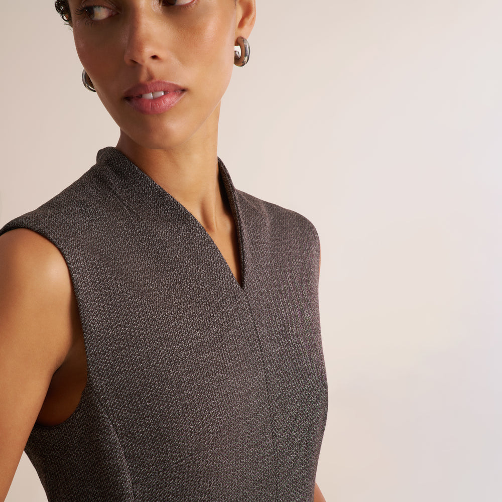 Close-up of a woman wearing a sleeveless textured wrap-style top in dark gray-brown, paired with small hoop earrings, against a plain light beige background.