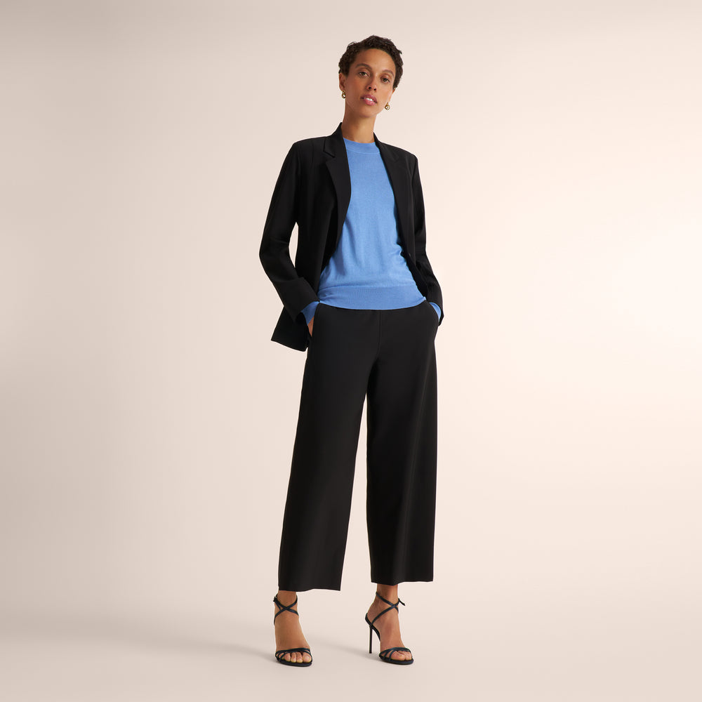 Woman wearing a black blazer and matching wide leg black pants with a blue splendor sweater underneath standing on a beige background