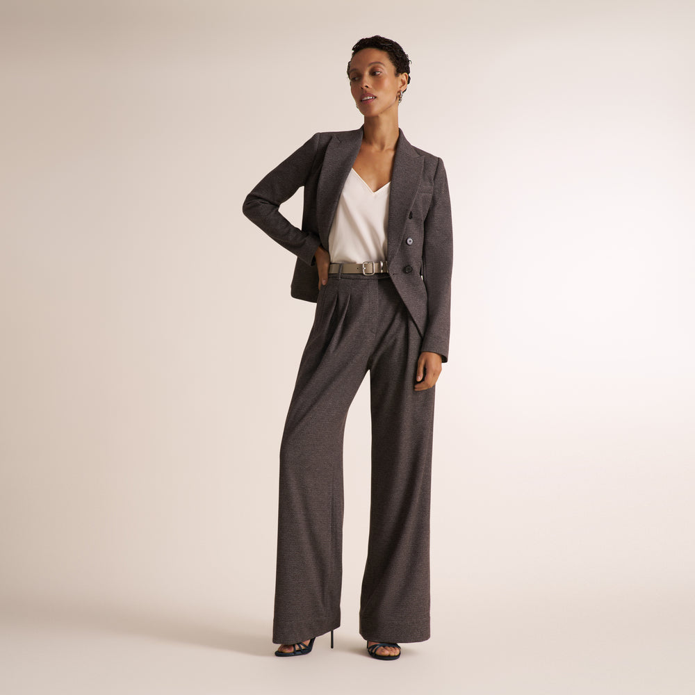 A model stands in a studio wearing a dark gray tailored pantsuit with a blazer and wide-leg trousers, styled with a white V-neck top, a thin metallic belt, and black high-heeled sandals against a light beige background.