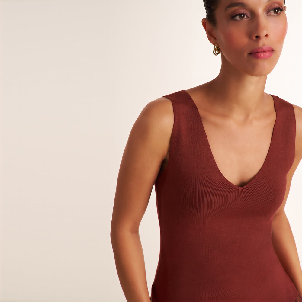 A close-up of a woman wearing a sleeveless deep V-neck knit top in a muted terracotta or rust red color, set against a plain light beige studio background.