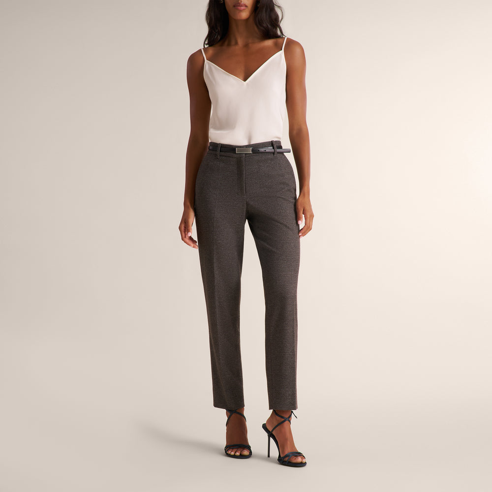 A model wearing a pair of dark gray textured straight-leg trousers with a black belt, paired with a white camisole and black strappy heels, against a solid light beige background.