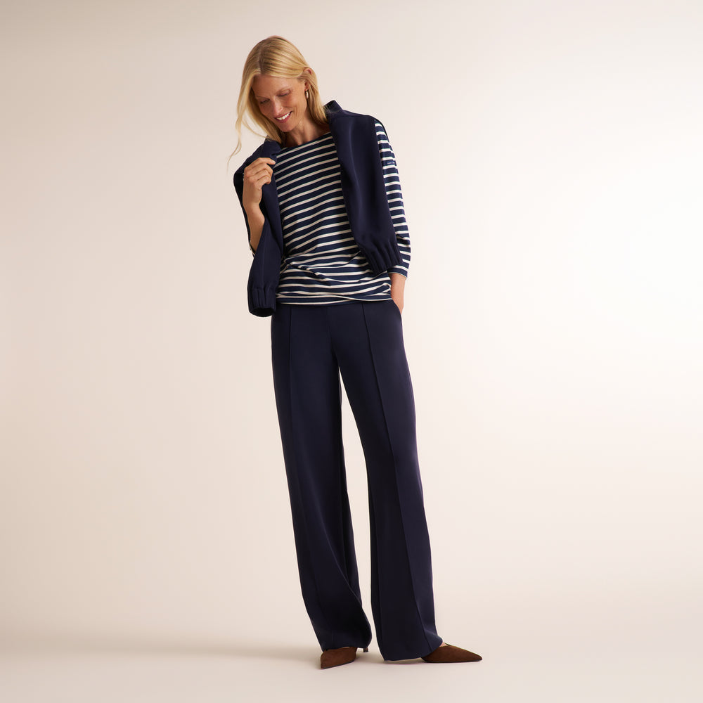 A woman modeling a navy blue sleeveless vest over a navy and white striped long-sleeve top, paired with navy blue wide-leg trousers and brown pointed-toe heels, against a solid light beige studio background.