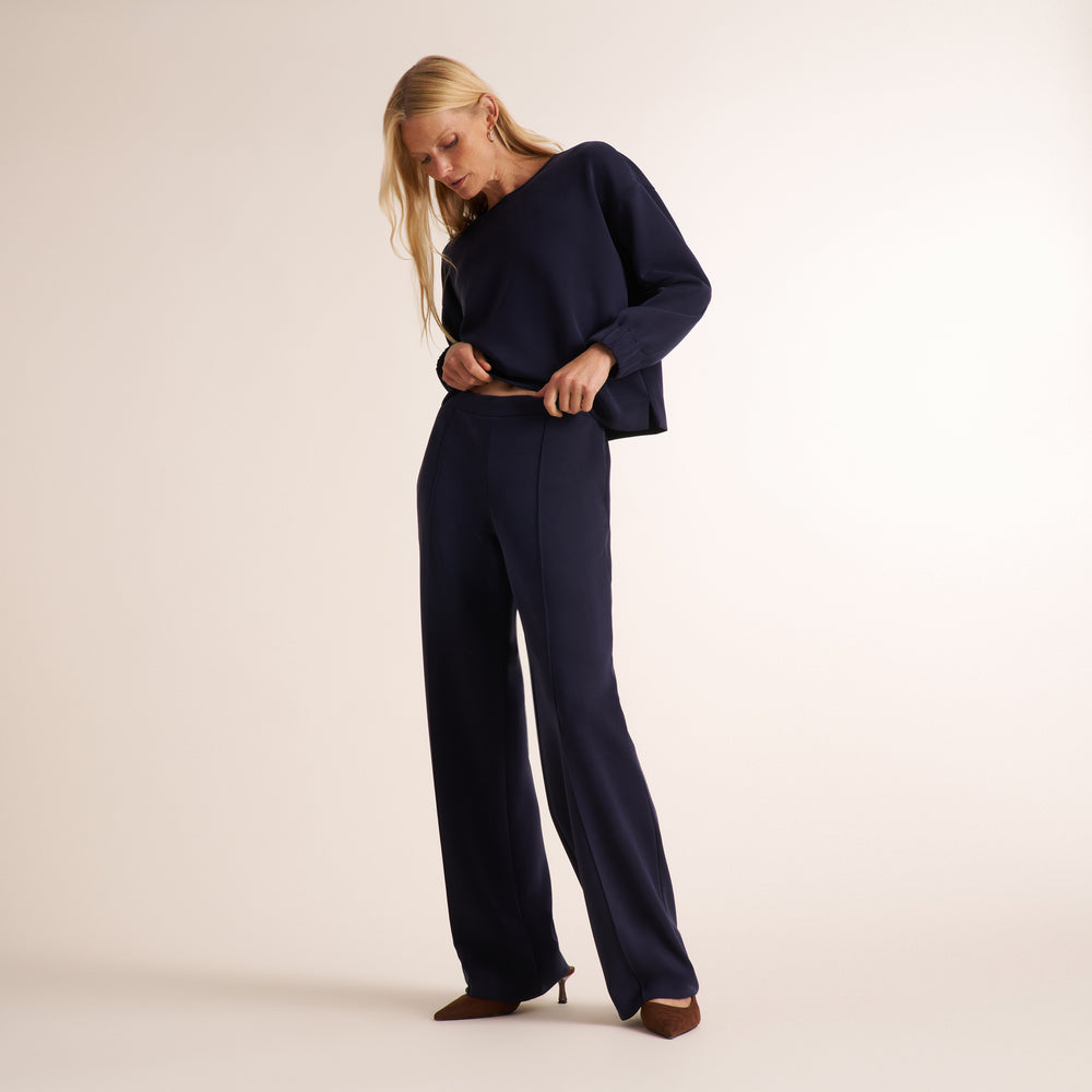 A woman modeling a navy blue two-piece outfit consisting of a long-sleeve asymmetrical top and matching wide-leg trousers, paired with brown pointed-toe heels, against a plain light beige studio background.