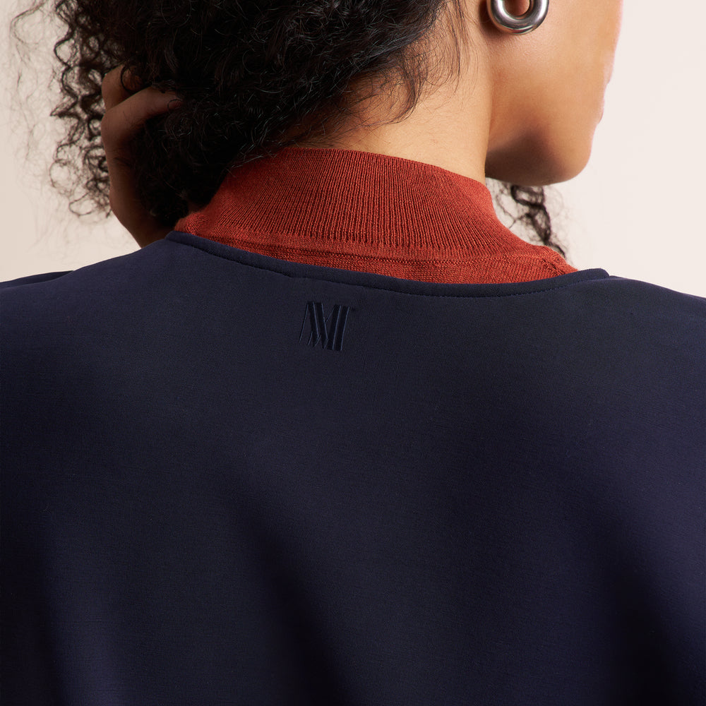 Close-up back view of a person wearing a navy blue sweatshirt with a rust-orange ribbed mock neck collar. A small tonal embroidered logo is visible at the center of the collar.