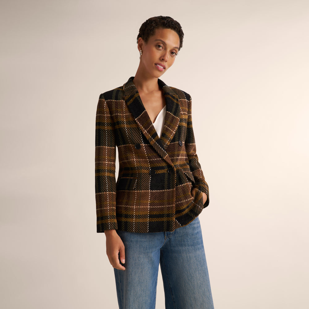 A woman modeling a brown and black plaid wrap blazer with a tie waist, paired with blue jeans, against a plain light beige studio background.