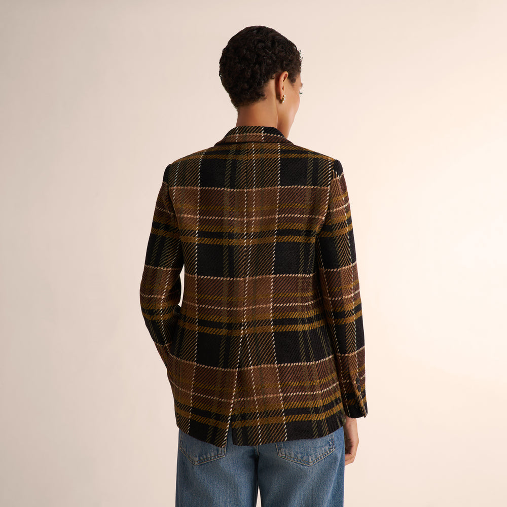 A person shown from the back wearing a brown, black, and beige plaid blazer with blue jeans, photographed against a plain light beige studio background.