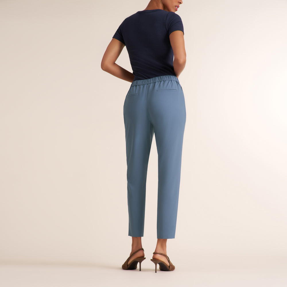 A woman shown from the back wearing light blue cropped trousers, a navy short-sleeve top, and brown heeled sandals against a plain beige studio background.