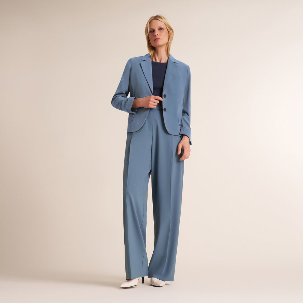 A woman stands against a plain warm beige studio background wearing a matching blue tailored set with a blazer and wide-leg trousers, layered over a dark top and white pointed shoes.