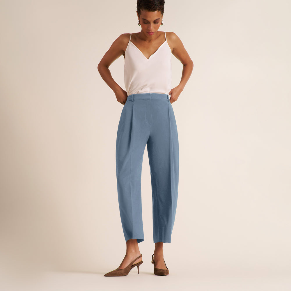 A model wearing a white camisole top and light blue cropped trousers, standing against a plain beige studio background.