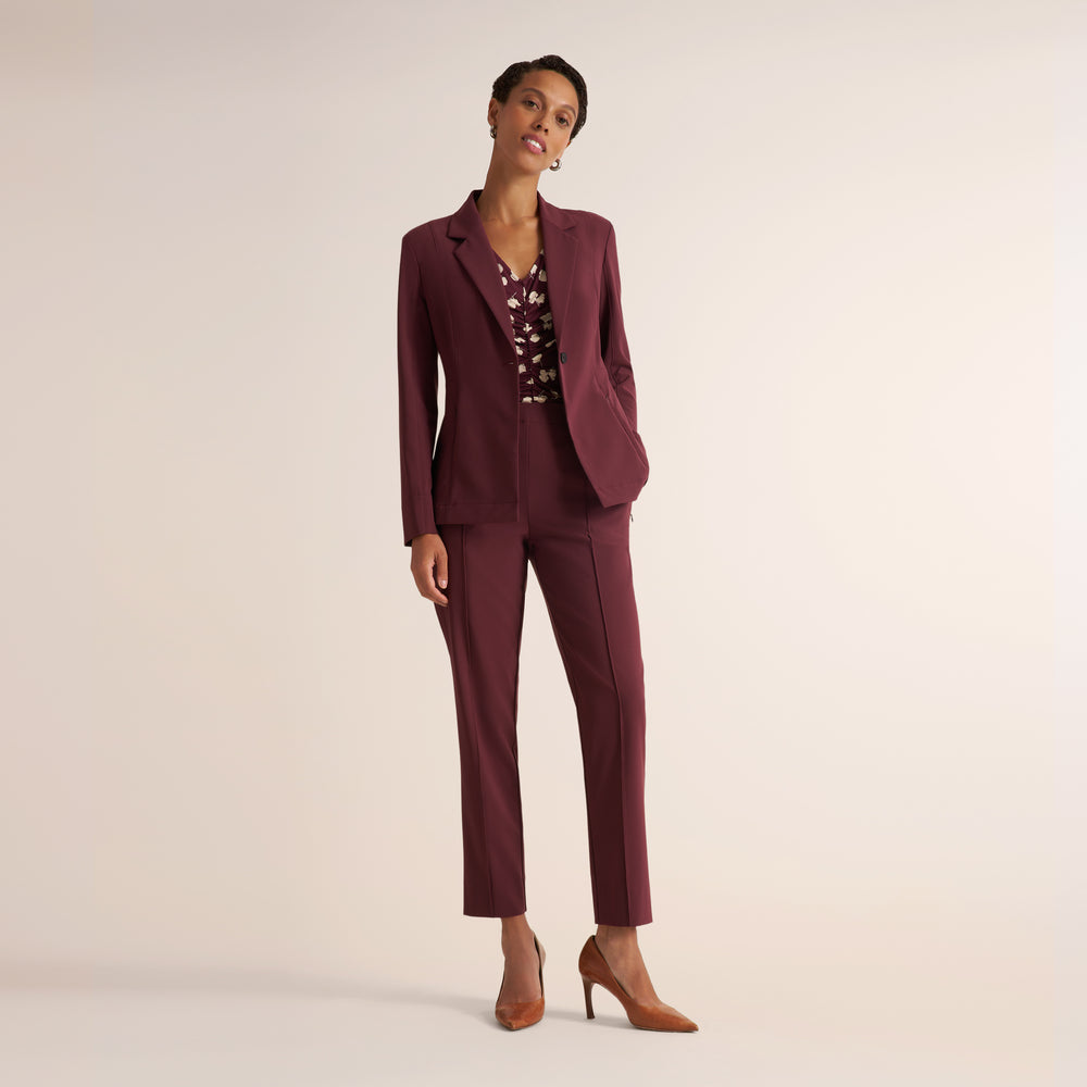A woman models a matching burgundy two-piece suit consisting of a tailored blazer and straight-leg cropped trousers, paired with a patterned top and tan heels, against a solid light beige studio background.