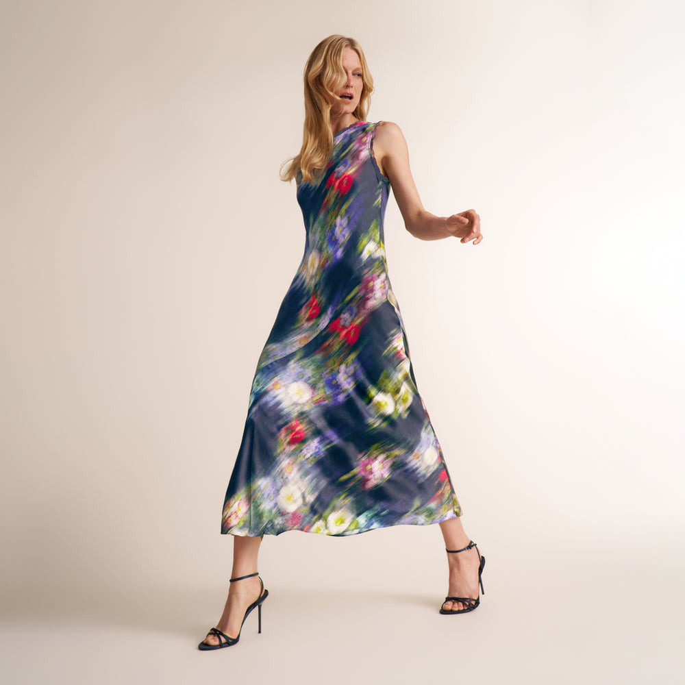 A woman modeling a sleeveless midi dress with a dark blue base and a colorful abstract floral print, paired with black strappy high-heeled sandals against a plain beige studio background.