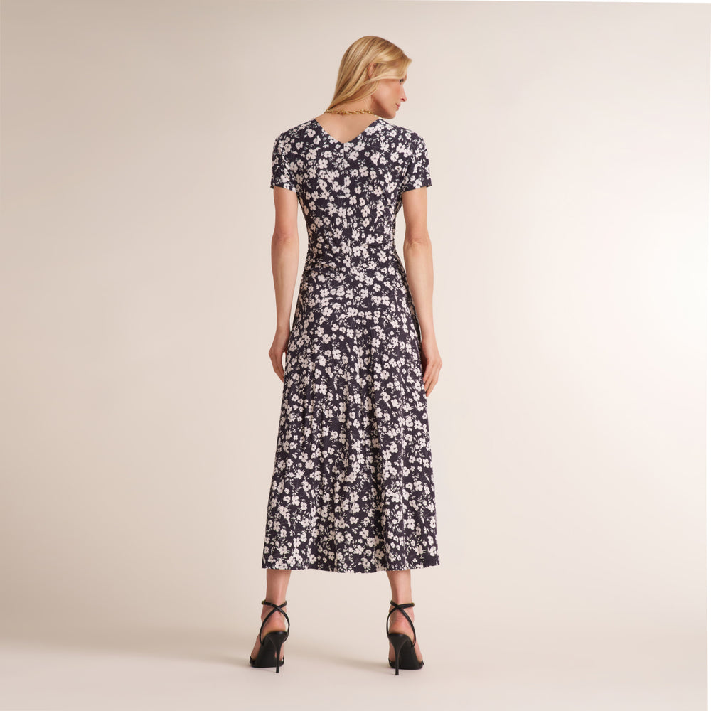 A woman stands with her back turned wearing a short-sleeved midi dress with a black and white floral pattern, paired with black high heels, against a solid beige studio background.