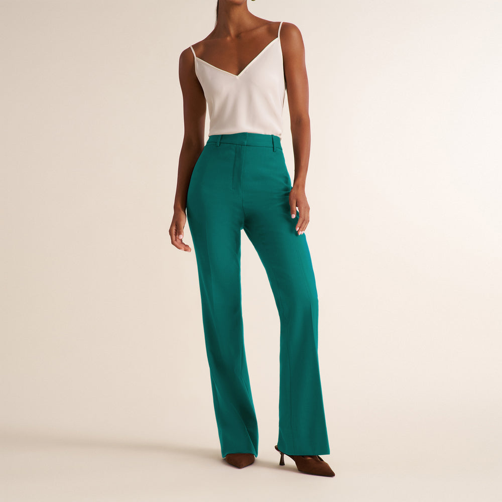 A model is shown from the neck down wearing high-waisted, wide-leg teal pants with a white camisole top and brown heels, set against a plain beige studio background.