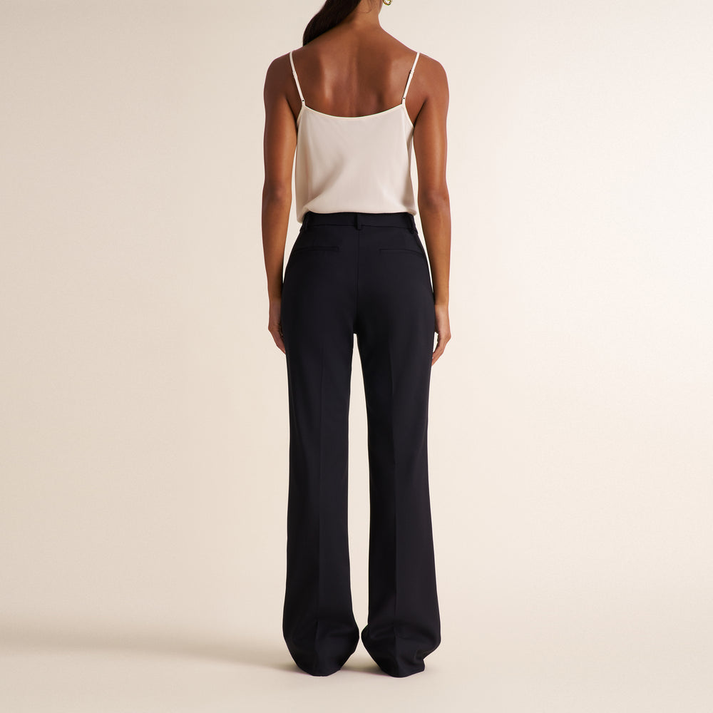 Woman wearing a black blazer, wide leg black suit pants, and a v-neck white tank top standing on a beige background
