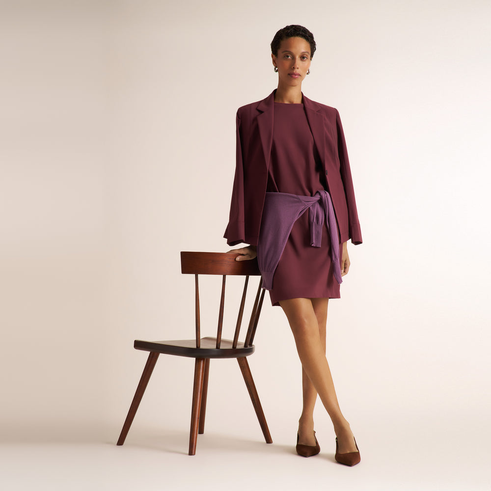 A model stands next to a wooden chair in a studio setting, wearing a matching two-piece outfit consisting of a deep plum blazer and a matching wrap-style mini dress with a waist tie.