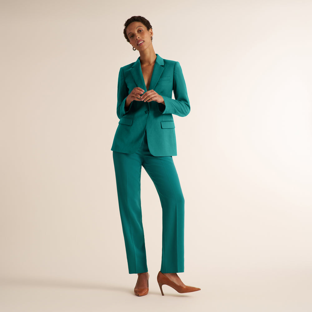 A woman modeling a teal green tailored pantsuit with a blazer and matching trousers, paired with brown pointed-toe heels, against a plain warm beige studio background.