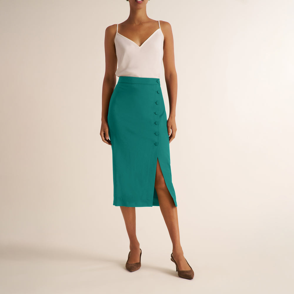 A model wearing a teal green high-waisted midi skirt with a button-down front and side slit, paired with a white camisole top and brown flat shoes, against a plain beige studio background.