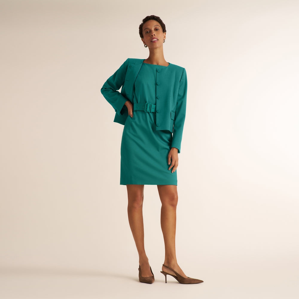 A woman models a teal green tailored dress with a matching jacket and belt, styled with brown pointed-toe heels against a plain beige studio background.